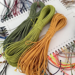 May include: Three bundles of elastic cord in olive green, lime green, and mustard yellow are arranged on a white display card. The card features numbered holes from 56 to 65, with small loops of various colored cords attached to each numbered position.