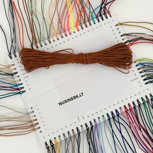 May include: A white rectangular display card featuring the text NUSINERK.LT, organized with numbered holes holding various colored elastic cords. A bundle of brown elastic cord is placed horizontally across the center of the display card.