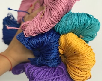 May include: A hand holds a collection of colorful macrame cords. The cords are in shades of pink, blue, teal, yellow, and purple. The cords are tightly wound and appear to be made of a smooth, synthetic material, perfect for crafting projects.