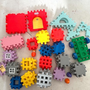May include: A collection of colorful plastic building blocks in various shapes and sizes, including squares, rectangles, and triangles. Some blocks have windows or doors, while others have a grid pattern. The blocks are arranged in a random pattern on a light gray surface.