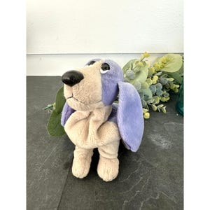 Peut inclure: Une peluche en forme de chien avec un poitrail et des pattes beige clair, ainsi que des oreilles, une tête et un dos de couleur lavande. Ce jouet en tissu doux possède de grands yeux sombres et brillants et un nez noir lisse et arrondi.
