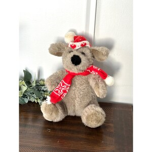 May include: A soft toy dog wearing a red and white knitted hat and scarf. The hat has white pom-poms and heart patterns. The scarf is red with white snowflake designs. The dog has a black nose and is seated.