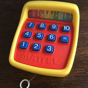 May include: A vintage Mattel toy calculator in red, yellow, and blue. The calculator features numbered buttons from 1 to 10, a row of numbers above the buttons, and the brand name "MATTEL" printed on the front. A white ring is attached to the toy.