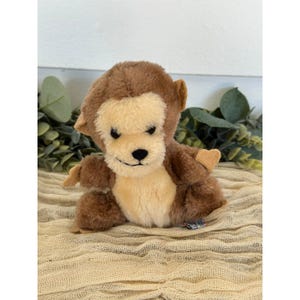 May include: A plush monkey toy with brown fur and a cream-colored face, chest, and hands. The monkey has a smiling expression and small, heart-shaped ears. It is sitting on a tan fabric surface.
