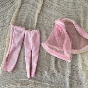 Op de afbeelding: Een poppenoutfit met een roze legging, bijpassende beenwarmers en een roze cape met een doorschijnende overlay en imitatiebont. De items zijn gerangschikt op een textiel met neutrale tint.