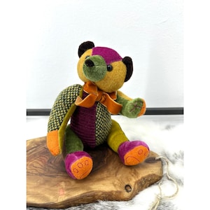 Peut inclure: Un ours en peluche fabriqué à partir de pièces de tissu aux textures variées dans des tons jaune moutarde, vert olive, violet profond et orange. L'ours porte un ruban en velours orange noué autour du cou et a des yeux en plastique noirs.