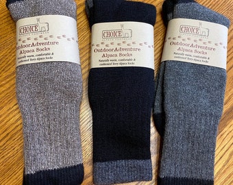 Puede incluir: Tres pares de calcetines de alpaca en tonos grises y negros, cada uno con puño y talón en contraste. Los calcetines están etiquetados como "Outdoor Adventure Alpaca Socks" con texto que los describe como naturalmente cálidos y cómodos.