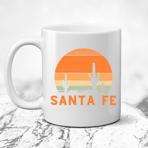 May include: White ceramic mug with a sunset graphic featuring orange, yellow, and green stripes and two cactus silhouettes. The text "SANTA FE" is printed in orange below the graphic.