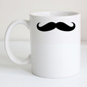 May include: White ceramic mug featuring a large black mustache design. The mug has a curved handle and a simple, clean aesthetic. A fun and quirky item for coffee or tea.
