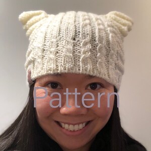 May include: A white knitted hat with ear flaps. The hat has a cable knit pattern. The word "Pattern" is visible in the image.