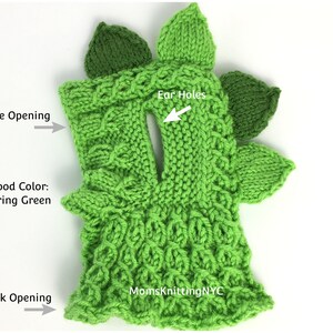 Op de afbeelding: Een lentegroene gebreide snood met een dinosaurusontwerp. De snood heeft een gezichtsopening, oor gaten en een nekopening. Het ontwerp bevat bladvormige accenten en de tekst "MomsKnittingNYC".