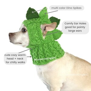 Op de afbeelding: Een groene gebreide dinosauruskap voor een hond, met veelkleurige stekels en oor gaten. De kap bedekt het hoofd en de nek en biedt warmte voor koude wandelingen. De tekst "MomsKnittingNYC" is zichtbaar.