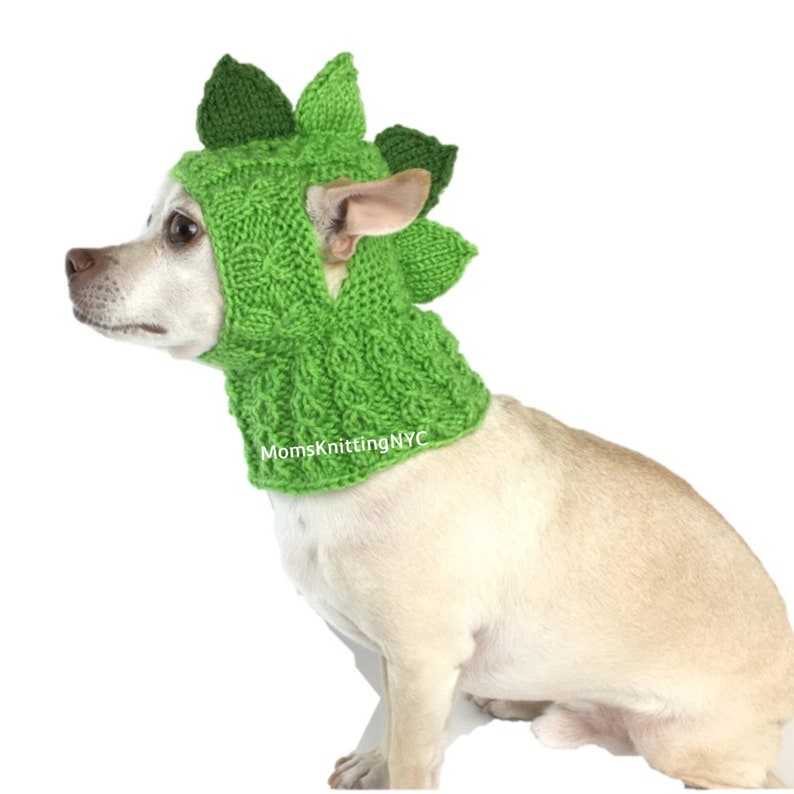 Op de afbeelding: Een kleine hond draagt een felgroene gebreide dinosauruskap. De kap heeft een textuurpatroon en stekelige details langs de bovenkant, die lijken op de rug van een dinosaurus. De tekst "MomsKnittingNYC" is zichtbaar op de kap.