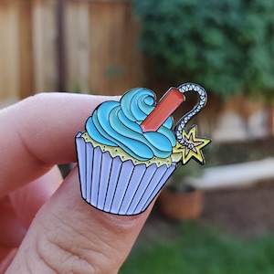 May include: A blue and purple cupcake with a red and black fuse and a yellow star on top. The cupcake is a pin.
