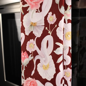 May include: A burgundy kitchen towel with a floral pattern. The design features large, light pink and white flowers with yellow centers and green leaves. The towel is hanging on a stainless steel oven.
