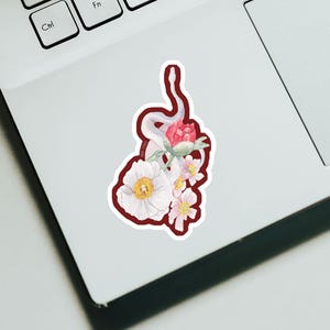 May include: A sticker featuring a white snake intertwined with a red rose and white and pink flowers. The sticker has a dark red border and is placed on a white laptop.