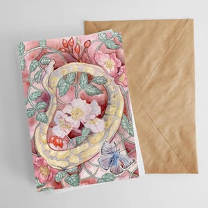 May include: A watercolor greeting card featuring a yellow snake coiled around pink flowers and green leaves. A blue butterfly is visible. The card is next to a brown envelope.
