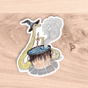 May include: A whimsical sticker featuring a yellow snake wearing a witch's hat, stirring a bubbling cauldron. The cauldron sits atop a fire, with steam rising. The sticker has a hand-drawn style and is set against a wooden background.