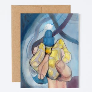 May include: Watercolor art print of a snake wearing a blue knit hat with a white pom-pom. The snake is yellow with a pattern of light yellow and gray spots. The background is blue with abstract shapes. The print is on a white card with a brown envelope.