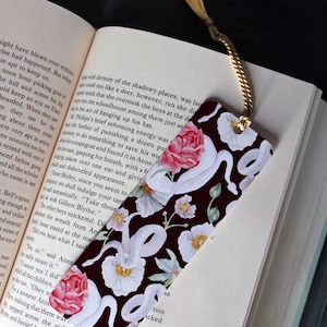 May include: A decorative bookmark with a dark burgundy background featuring a white snake and floral pattern. The bookmark has a gold tassel attached to the top and is placed in an open book.