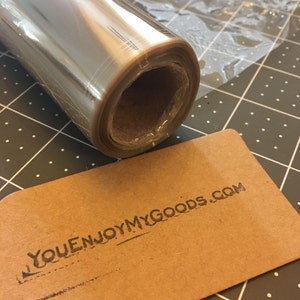 May include: A roll of clear cellophane wrapping paper on a cardboard tube. The paper is rolled up tightly and the end of the roll is visible. The paper is transparent and has a slight sheen. The roll is sitting on a gray and white checkered surface. A brown tag with the text "YOUENJOYMYGOODS.COM" is next to the roll.