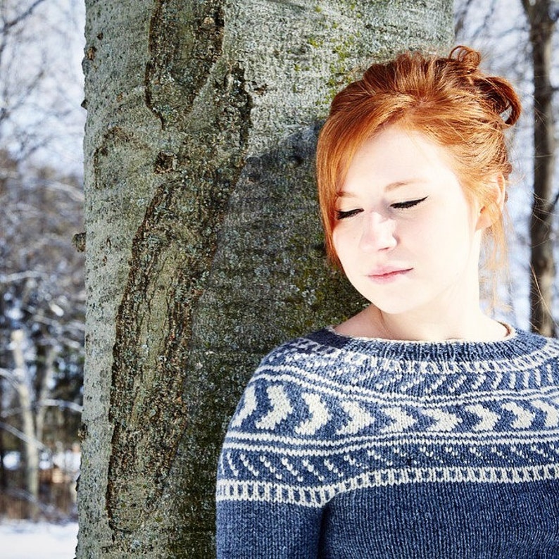 K&ouml;nnte beinhalten: Eine Frau mit roten Haaren tr&auml;gt einen blau-wei&szlig;en Pullover mit Muster und steht neben einem Baumstamm.