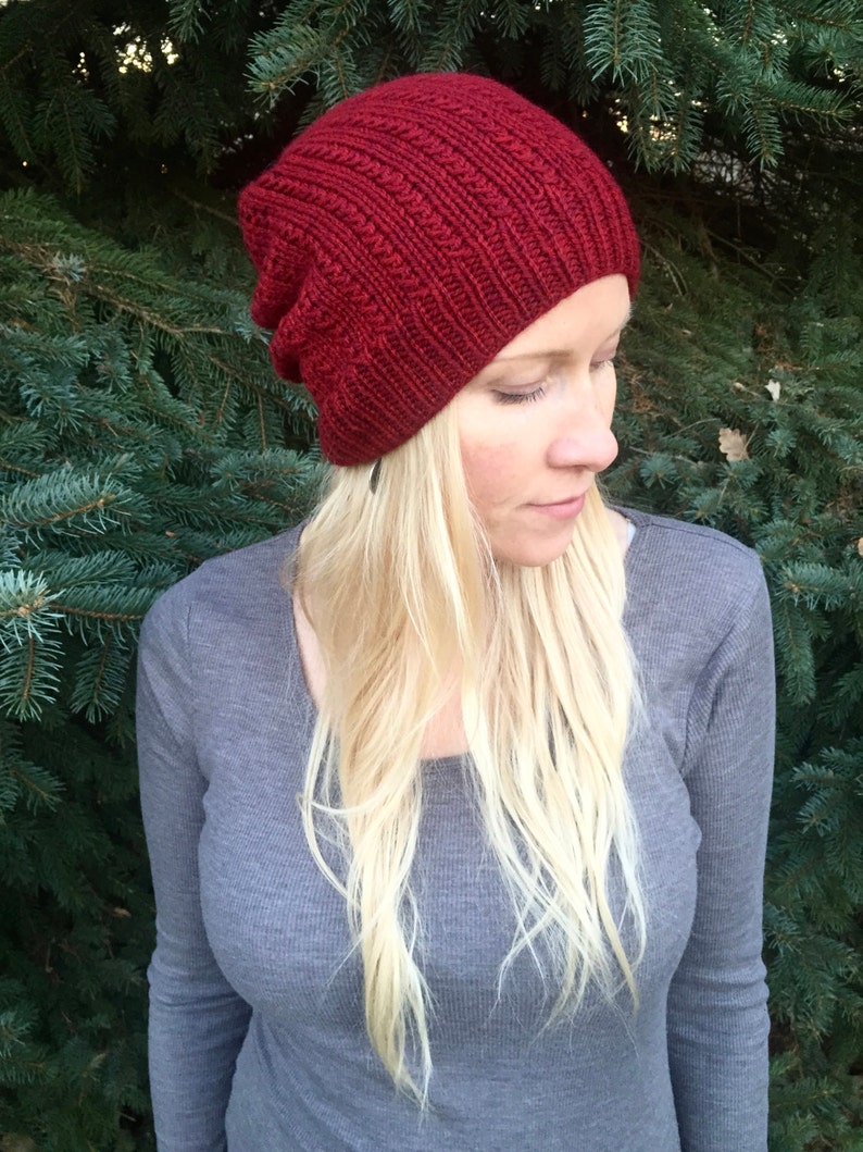 May include: A close-up of a woman wearing a red knitted beanie hat. The hat has a slouchy fit and a ribbed texture. The woman has long blonde hair and is wearing a gray long-sleeved shirt.