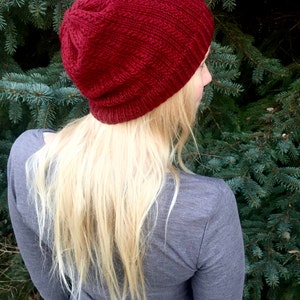 May include: A close-up of a woman wearing a red knitted beanie hat. The hat is slouchy and has a ribbed texture. The woman has long blonde hair and is wearing a gray shirt.