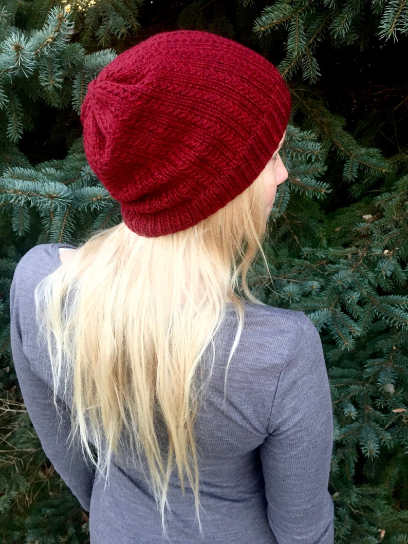 May include: A close-up of a woman wearing a red knitted beanie hat. The hat is slouchy and has a ribbed texture. The woman has long blonde hair and is wearing a gray shirt.