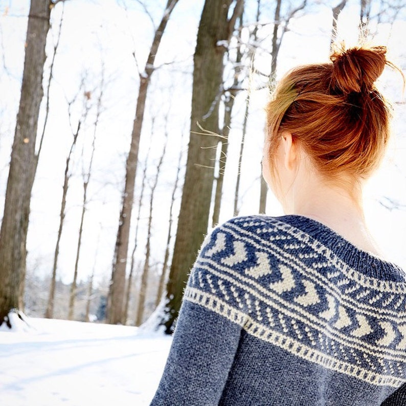 K&ouml;nnte beinhalten: Eine Frau mit roten Haaren in einem Dutt tr&auml;gt einen blau-wei&szlig;en Strickpullover mit geometrischem Muster. Sie steht in einem schneebedeckten Wald mit B&auml;umen im Hintergrund.