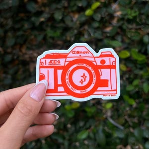 May include: A sticker featuring a red and pink Canon AE-1 Program camera design. The sticker includes the Canon logo, lens details, and the name "Maiya Buck". The camera design is outlined in white.
