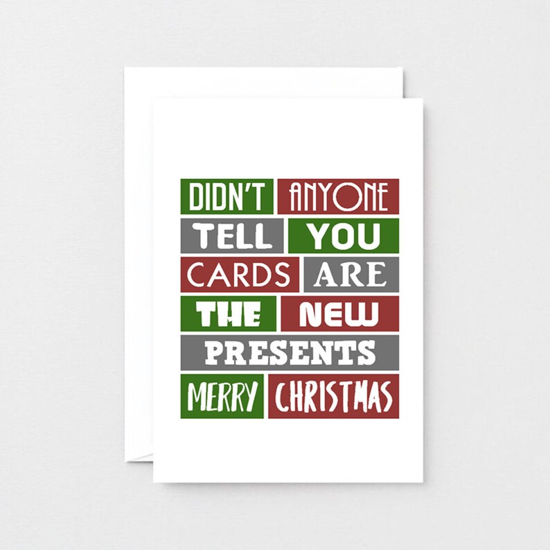 Pu&ograve; includere: Un biglietto d'auguri bianco con una griglia di rettangoli rossi, verdi e grigi. Il testo recita: DIDN'T ANYONE TELL YOU CARDS ARE THE NEW PRESENTS MERRY CHRISTMAS. Il design utilizza un mix di caratteri bianchi con e senza grazie.