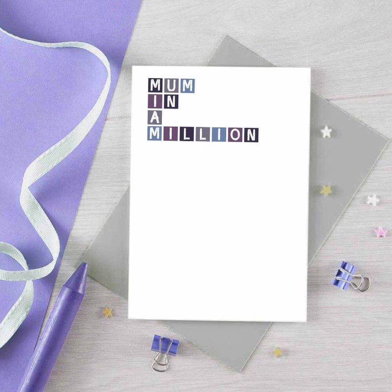 May include: A white card with the words "MUM IN A MILLION" spelled out in colorful squares. The card is on a gray surface with a purple ribbon and a purple crayon.