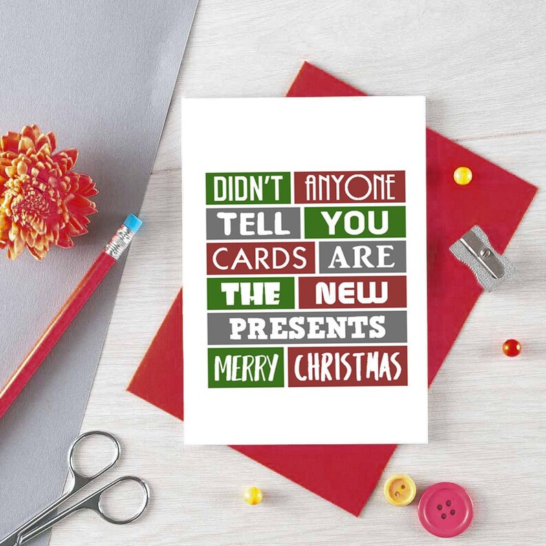 Pu&ograve; includere: Un biglietto d'auguri rettangolare bianco con il testo DIDN'T ANYONE TELL YOU CARDS ARE THE NEW PRESENTS MERRY CHRISTMAS in blocchi rettangolari alternati di colore rosso, verde e grigio. Il biglietto &egrave; su uno sfondo di carta rossa.