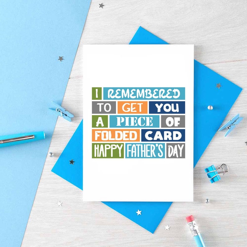 Pode incluir: Um cart&atilde;o branco com uma mensagem colorida que diz "Eu me lembrei de pegar um peda&ccedil;o de cart&atilde;o dobrado Feliz Dia dos Pais". O cart&atilde;o est&aacute; em um envelope azul e uma superf&iacute;cie de madeira.