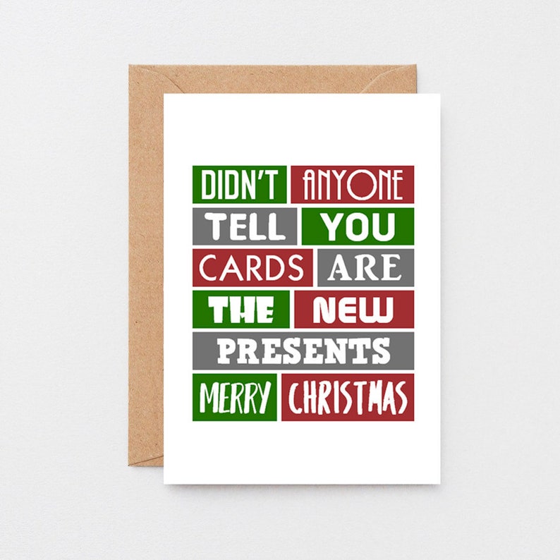 Pu&ograve; includere: Un biglietto d'auguri bianco con una busta marrone che presenta il testo DIDN'T ANYONE TELL YOU CARDS ARE THE NEW PRESENTS MERRY CHRISTMAS disposto in una griglia di blocchi rettangolari rossi, verdi e grigi con scritte bianche sul davanti.