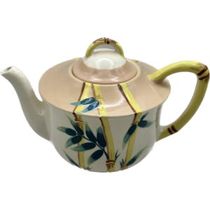 Puede incluir: Tetera de cerámica color crema con tapa y asa. La tetera presenta una tapa beige con rayas amarillas y un asa pequeña. El cuerpo está decorado con hojas de bambú verdes y tallos de bambú amarillos. El pico y el asa son amarillos.
