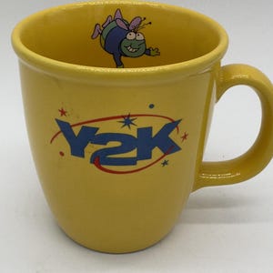 May include: Yellow ceramic mug with a curved handle. The mug features the blue text "Y2K" with star accents and a cartoon bug inside. The mug is a bright yellow color.