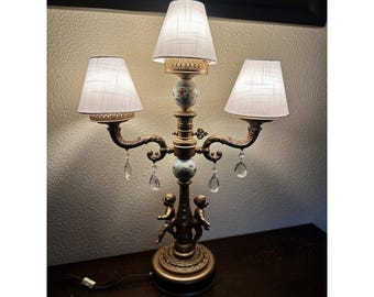 Może przedstawiać: Trójramienna lampa stołowa z podstawą z pozłacanego metalu i białymi akcentami z porcelany. Lampa posiada trzy białe abażury z tkaniny i kryształowe akcenty. Podstawa jest ozdobiona figurkami cherubów.