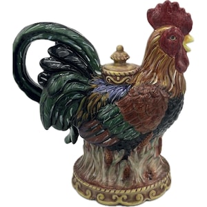 Pode incluir: Bule de cerâmica em forma de galo com um design detalhado. O bule apresenta uma crista vermelha, penas marrons e douradas e uma cauda verde. A base é um design de tronco de árvore com uma borda decorativa.