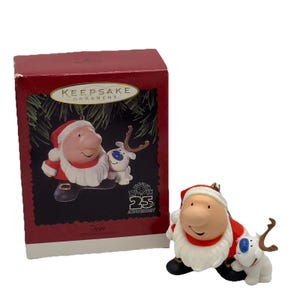 May include: A Hallmark Keepsake Christmas ornament featuring a cartoon Santa Claus and a reindeer. The Santa figure wears a red suit and hat with a white beard. The reindeer has brown antlers and a blue eye. The box reads "Keepsake Ornament".