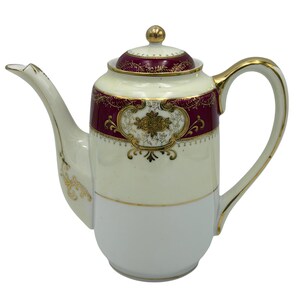 May include: An antique porcelain coffee pot with a cream-colored body and a white base. The pot features a burgundy band with gold floral detailing and a gold handle and spout. A gold finial tops the lid.