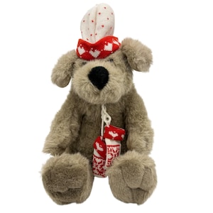 May include: A soft toy dog with a brown body, black nose, and floppy ears. It is wearing a red and white heart-patterned hat and scarf, and holds two miniature red and white heart-patterned stockings. The toy is designed for decorative purposes.