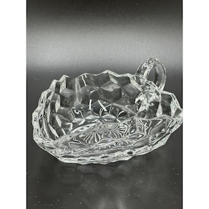 May include: A clear, faceted glass dish with a handle. The dish has a diamond pattern on the sides and a floral design in the center. The handle is a small, rounded loop. The dish is likely for serving or display.
