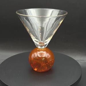 May include: A clear martini glass with a gold rim sits atop an orange, spherical base. The base has a bubbly texture. The glass has a classic cone shape, perfect for cocktails.