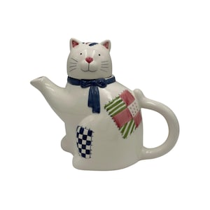 Pode incluir: Bule de chá de cerâmica branca em forma de gato com laço azul e desenhos de patchwork. O bule tem um bico, uma alça e uma tampa na cabeça do gato. O patchwork inclui padrões xadrez rosa, verde e azul.