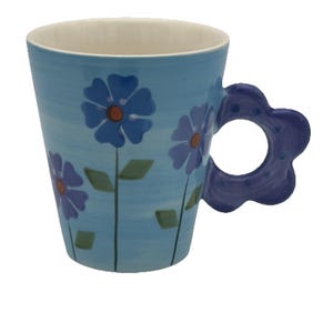May include: A light blue ceramic mug with a white interior and a flower-shaped handle. The mug is decorated with blue and purple flowers with orange centers and green leaves. The mug is approximately 4 inches tall.