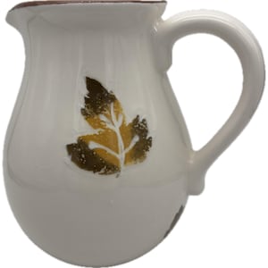May include: Cream-colored ceramic pitcher with a brown rim and handle. The pitcher features a leaf design in shades of brown and yellow. The pitcher is approximately 7 inches tall.