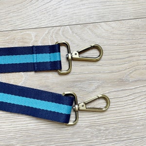 May include: Two navy blue and turquoise striped straps with gold metal clasps. The straps are on a light wood surface.