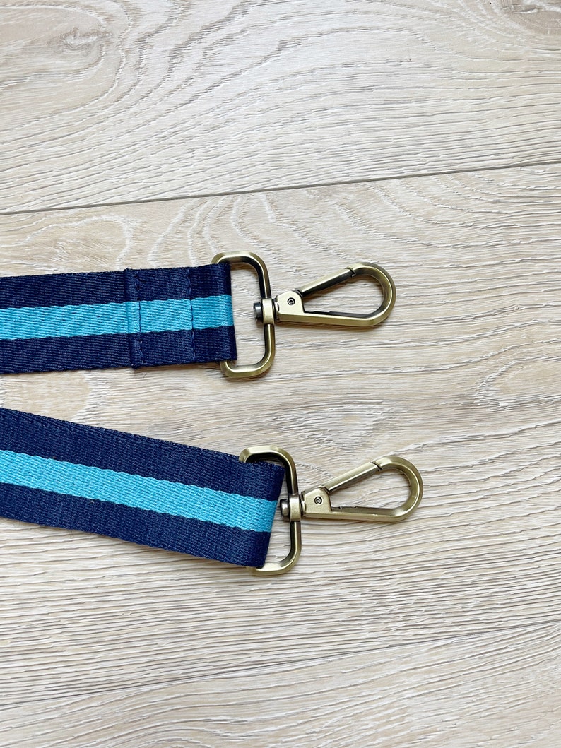 May include: Two navy blue and turquoise striped straps with gold metal clasps. The straps are on a light wood surface.